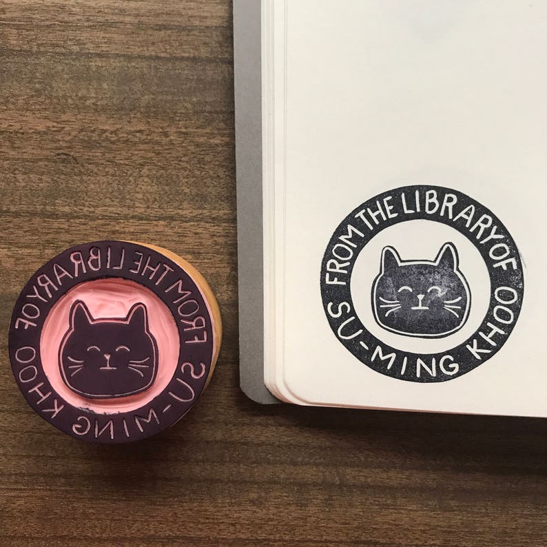 May include: Two round wooden stamps with a black cat design. The stamps say "Greetings from the Library of Su-Ming Khoo".