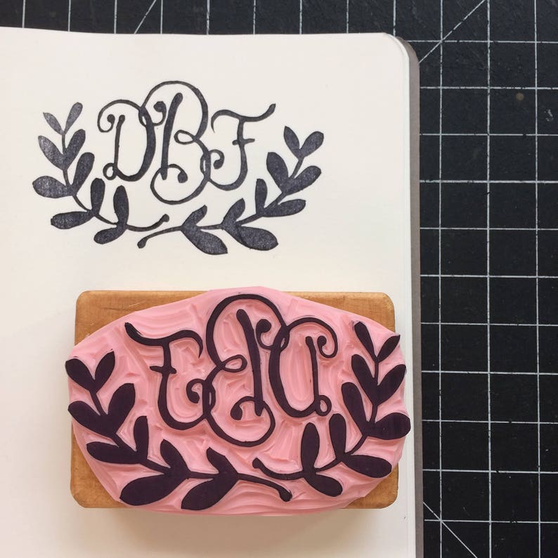 May include: A wooden block with a pink rubber stamp featuring a black monogram with the letters "EBU" surrounded by a wreath of leaves.
