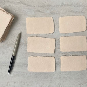 May include: Six blank, off-white, rectangular pieces of handmade paper with deckled edges. The paper is arranged in two rows of three. A silver pen is lying on a gray surface.
