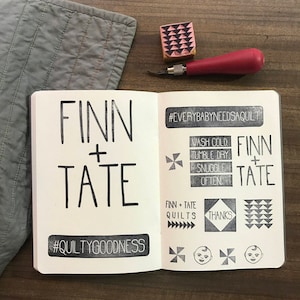 May include: Open notebook with stamped text reading "FINN + TATE" and "#QUILTYGOODNESS". Additional stamps include "#EVERYBABYNEEDSAQUILT" and care instructions. A gray quilted fabric and a carving tool are also visible.