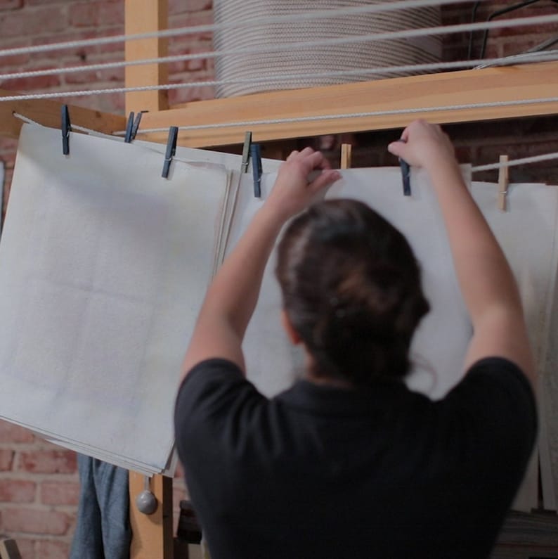 May include: A person hanging up white sheets of paper on a clothesline with clothespins. The sheets are hanging in a room with brick walls and a wooden shelf.