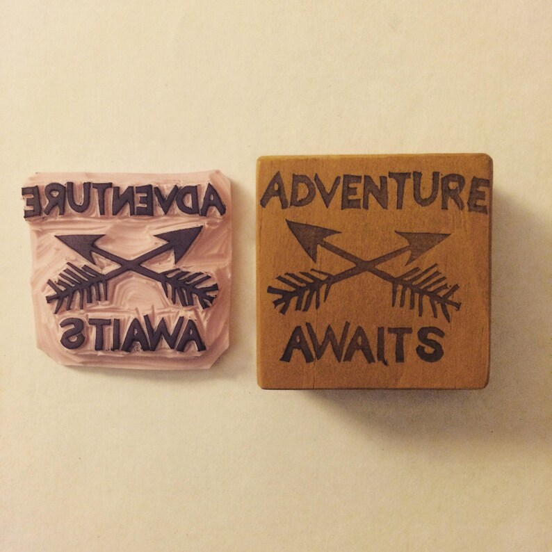 May include: A wooden stamp with the words "ADVENTURE AWAITS" and two crossed arrows. Next to it is the rubber stamp with the reversed image. The stamp is brown, and the rubber stamp is pink.