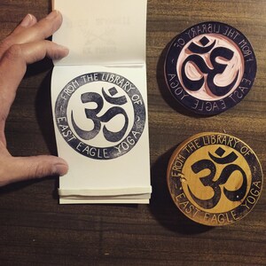 May include: Three wooden stamps with the Om symbol and the text "From the Library of East Eagle Yoga". The stamps are in different colors: black, brown, and pink.