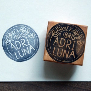 May include: A close-up of a wooden stamp with a black ink pad. The stamp features a circular design with the text "Ex Libris Adri Luna" in a white font. The design also includes a wreath of leaves around the text.