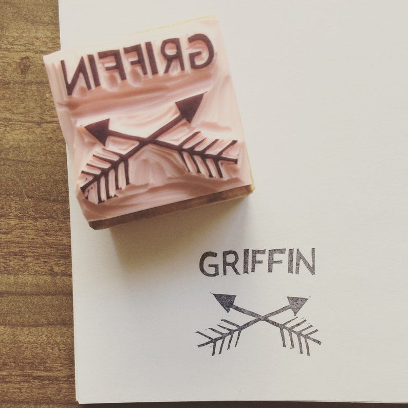 May include: A pink rubber stamp with the word "GRIFFIN" in dark brown letters and two crossed arrows. The stamp is pressed onto white paper, leaving a matching imprint of the design.