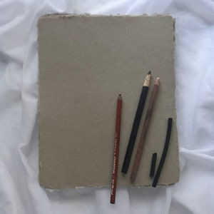 May include: Four drawing pencils and a stick of charcoal on a light gray textured paper surface. The pencils are brown, black, and red. The charcoal is black. The pencils are arranged vertically and the charcoal is horizontal.