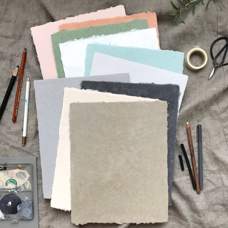 May include: A stack of eight different colored sheets of textured paper, with a variety of drawing pencils and charcoal sticks on a linen fabric background.