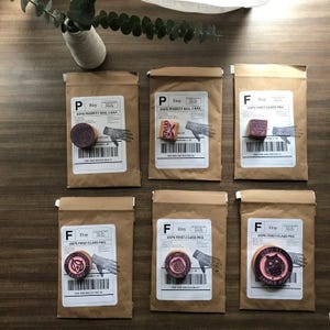 May include: Six brown paper packages with stamps and labels. Each package has a different stamp design, including a hand, an owl, and various circular designs. The packages are arranged on a wooden surface, with a vase of greenery in the background.