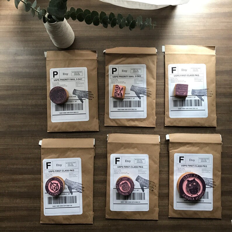 May include: Six brown paper packages with stamps and labels. Each package has a different stamp design, including a hand, an owl, and various circular designs. The packages are arranged on a wooden surface, with a vase of greenery in the background.