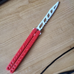 May include: A red and white butterfly knife, also known as a balisong, is displayed on a wooden surface. The handle is red, and the blade is white with a series of cut-out shapes. The knife is in the open position.