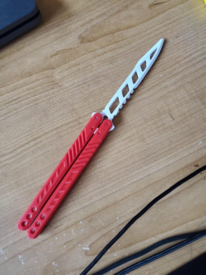 May include: A red and white butterfly knife, also known as a balisong, is displayed on a wooden surface. The handle is red, and the blade is white with a series of cut-out shapes. The knife is in the open position.