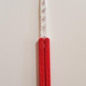 May include: A red and white butterfly knife. The blade is white with a series of cut-out shapes. The handles are red with diagonal grooves and three holes near the bottom. The knife is shown against a white background.