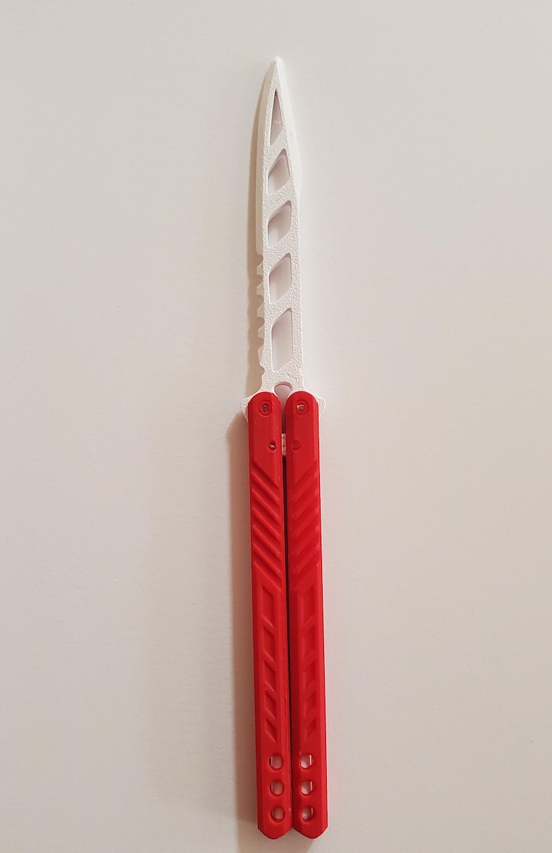 May include: A red and white butterfly knife. The blade is white with a series of cut-out shapes. The handles are red with diagonal grooves and three holes near the bottom. The knife is shown against a white background.
