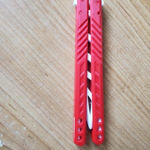 May include: A red and white butterfly knife, also known as a balisong, is shown. The handles are red with a ridged design, and the blade is white. The knife is closed and resting on a wooden surface.