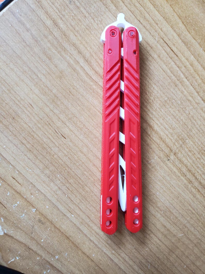 May include: A red and white butterfly knife, also known as a balisong, is shown. The handles are red with a ridged design, and the blade is white. The knife is closed and resting on a wooden surface.