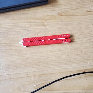 May include: A red and white butterfly knife, closed and resting on a wooden surface. The knife's handles are bright red with white accents and circular details. The blade is folded inside the handles.