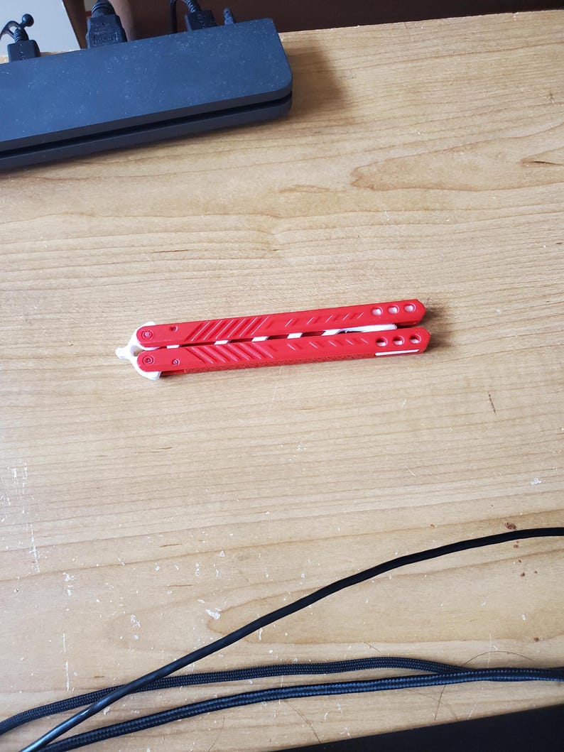 May include: A red and white butterfly knife, closed and resting on a wooden surface. The knife's handles are bright red with white accents and circular details. The blade is folded inside the handles.