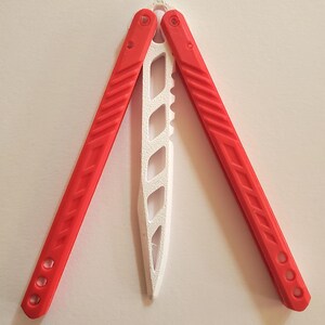 May include: A red and white butterfly knife, also known as a balisong, is displayed open against a white background. The handles are red with a textured design, and the blade is white with a series of cut-out shapes.