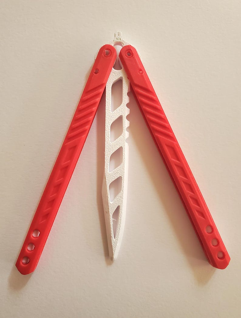 May include: A red and white butterfly knife, also known as a balisong, is displayed open against a white background. The handles are red with a textured design, and the blade is white with a series of cut-out shapes.