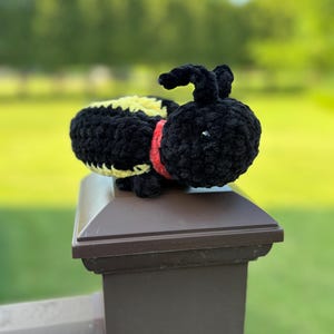 May include: A plush toy shaped like a bee, crafted from soft black and yellow yarn. The bee features a rounded black head with small antennae, a red collar, and a striped black and yellow body. The texture is fuzzy and soft to the touch.