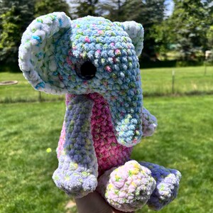 May include: A plush elephant toy crafted from variegated yarn in shades of light blue, pink, white, and yellow. The stuffed animal features large ears, a trunk, and dark, round plastic eyes, with a textured, soft appearance throughout its body.