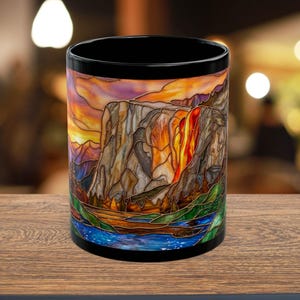 May include: Black ceramic mug featuring a stained-glass design of a mountain landscape with a waterfall. The artwork includes a sunset with orange, yellow, and purple hues. The base of the mug has a blue river and green trees.