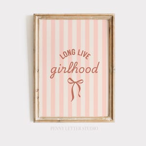 May include: A framed print featuring a pink and cream striped pattern. The words "LONG LIVE girlhood" are displayed in a cursive font, with a bow illustration beneath. The frame has a light brown finish.