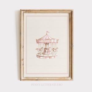May include: A framed print with a watercolour illustration of a carousel. The carousel has a pink and white striped canopy and several horses. The print is set against a cream background and framed in a gold-toned wooden frame. The text "PENNY LETTER STUDIO" is at the bottom.