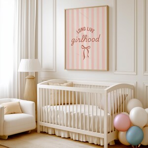 A white crib with a pink and white striped poster above it, a white chair, and a bunch of balloons in the corner of the room.