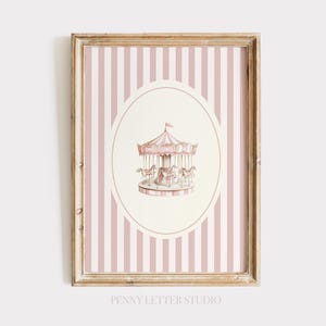 May include: A framed print with a watercolour illustration of a carousel. The carousel is pink and white, set within an oval against a background of vertical pink and white stripes. The frame is light brown. The text "PENNY LETTER STUDIO" is at the bottom of the print.