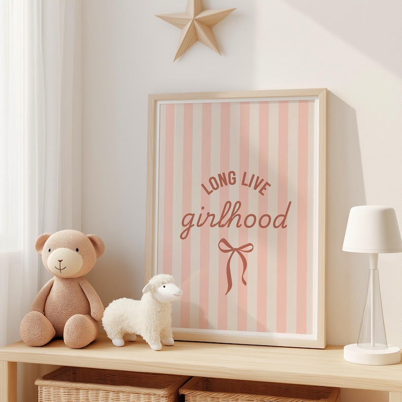 a wooden shelf with a framed poster, a teddy bear, and a lamp. The poster has a pink and white striped background and the text "Long Live Girlhood" written in a cursive font.