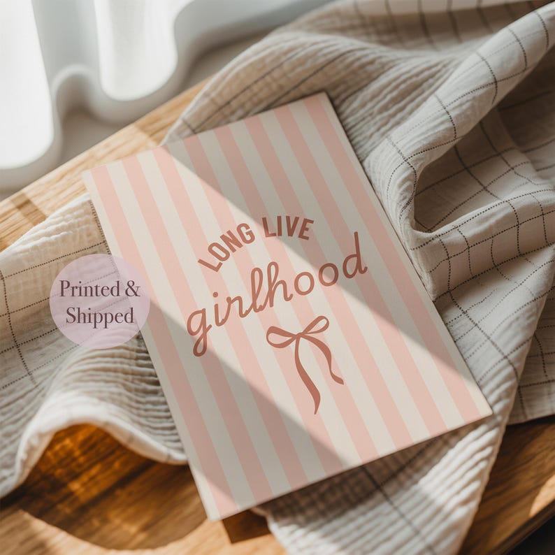 A printed and shipped card with the text "LONG LIVE GIRLHOOD" printed on it, placed on a wooden surface next to a striped towel.
