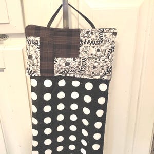 May include: A hanging kitchen towel with a black and white polka dot design. The top section has a patchwork design with brown plaid and floral patterns. A black strap is attached for hanging.