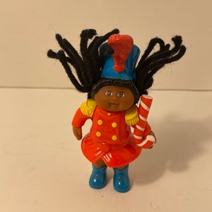 May include: A black Cabbage Patch Kid doll dressed as a toy soldier. The doll has black yarn hair, a red and gold uniform, a blue hat with a red plume, and is holding a candy cane.