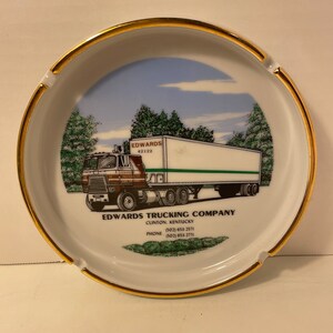 May include: White ceramic ashtray with a gold rim. The ashtray features a colorful illustration of a semi-truck with the text "EDWARDS TRUCKING COMPANY, CLINTON, KENTUCKY (502) 653-2571 PHONE (502) 653-2771".