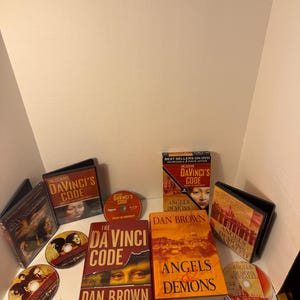 May include: A collection of books and DVDs, including "The Da Vinci Code" and "Angels & Demons" by Dan Brown. The items are arranged on a white surface, with the book covers and DVD cases visible. The color scheme is dominated by reds, oranges, and browns.