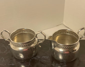 Puede incluir: Un juego de jarra de crema y azucarero de plata con un diseño de cuerda decorativo alrededor del borde.