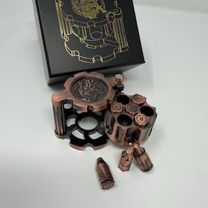 May include: A copper-coloured, steampunk-style ashtray set with a revolver cylinder design. The set includes a round ashtray, a cylinder with numbered compartments, and several bullet-shaped containers. A black box with gold-coloured gear artwork is in the background.