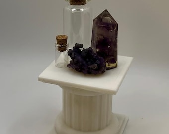 Puede incluir: Un pedestal estriado blanco sostiene dos frascos de vidrio transparente con tapones de corcho, una punta de cristal de amatista de color púrpura oscuro y un pequeño racimo de minerales botrioidales púrpura sobre la superficie plana.