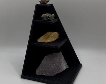 Könnte beinhalten: Ein schwarzes, dreieckiges Ausstellungsregal mit vier Ebenen, die jeweils ein anderes Mineralexemplar enthalten. Zu den Mineralien gehören ein dunkelbrauner Felsen, ein Pyritcluster, ein gelb-oranger Stein und ein großer, violetter Kristall.