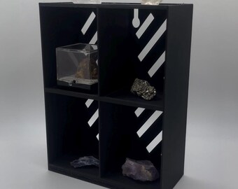 Könnte beinhalten: Ein schwarzes Regal mit vier Fächern und weißen diagonalen Streifen. Das Regal enthält verschiedene Gesteine und Mineralien, darunter Bergkristall, Pyrit und Amethyst. Ein Fach enthält eine kleine, transparente Vitrine.