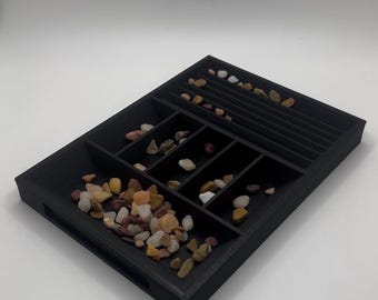 Könnte beinhalten: Ein schwarzer, rechteckiger Organizer mit mehreren Fächern, gefüllt mit kleinen, bunten Kieselsteinen. Der Organizer hat eine Reihe paralleler Rillen auf einer Seite und ist für das Halten und Präsentieren kleiner Gegenstände konzipiert.