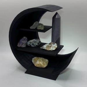 May include: A black crescent moon-shaped shelf with three horizontal tiers. The shelf holds various raw and polished mineral specimens, including a tall dark crystal point, a purple amethyst cluster, and several smooth, multi-colored stones.