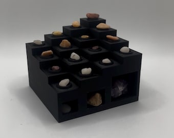 Könnte beinhalten: Ein schwarzer, abgestufter Präsentationsständer mit mehreren quadratischen Öffnungen, die jeweils einen kleinen Stein enthalten. Die Steine variieren in der Farbe, darunter Weiß, Beige und Braun. Der Ständer steht auf weißem Hintergrund.