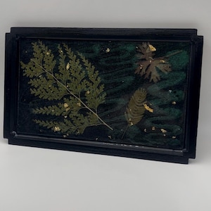 May include: A rectangular black framed resin art piece. The artwork features preserved green fern leaves and other foliage with gold leaf accents, set against a dark green background. The frame is a simple, modern design.