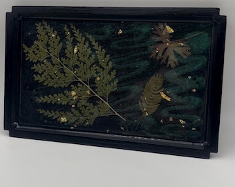Könnte beinhalten: Ein rechteckiges Kunstwerk aus Harz mit schwarzem Rahmen. Das Kunstwerk zeigt konservierte grüne Farnblätter und anderes Laub mit Goldblattakzenten vor einem dunkelgrünen Hintergrund. Der Rahmen hat ein schlichtes, modernes Design.