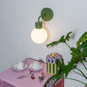 May include: A green wall-mounted light fixture with a round, white bulb. A pink and red side table holds two white teacups on saucers, spoons, a striped container, and a toy car. A green plant is in the background.