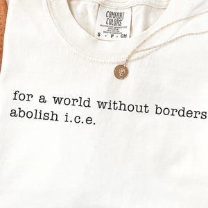 May include: Ivory-colored t-shirt with the text "for a world without borders abolish i.c.e." in black. A gold necklace with a pendant is draped across the shirt. The shirt is folded and placed on a white surface.
