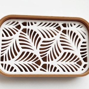 May include: Rectangular decorative tray with a white, leaf-patterned insert and a brown border. The tray's design features a repeating pattern of stylized leaves, creating an openwork effect. The tray is likely made of plastic or wood.