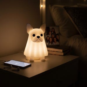 May include: A white, dog-shaped night light with a soft glow, sitting on a nightstand. The lamp has black eyes and a black nose. A smartphone with earbuds is next to the lamp. A framed photo and string lights are in the background.